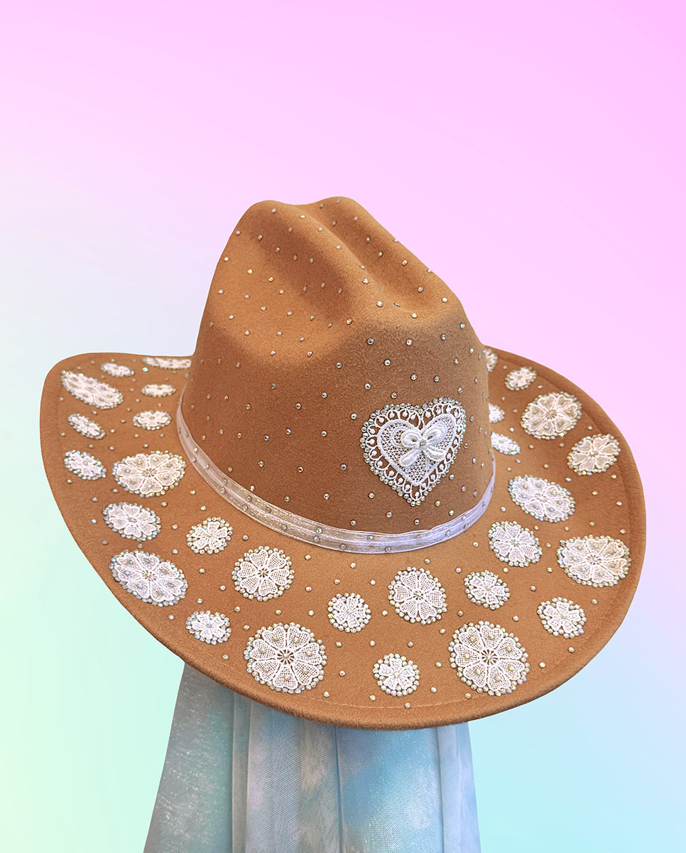 Brown cowboy hat with white lace heart and rhinestone embellishments on brim and crown.