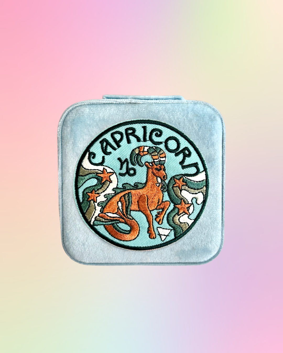 Capricorn zodiac sign design on a square, light blue, jewelry case against a pastel gradient background.