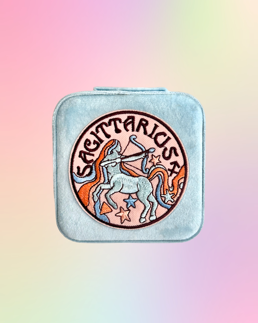 Sagittarius zodiac sign design on a square, light blue, jewelry case against a pastel gradient background.