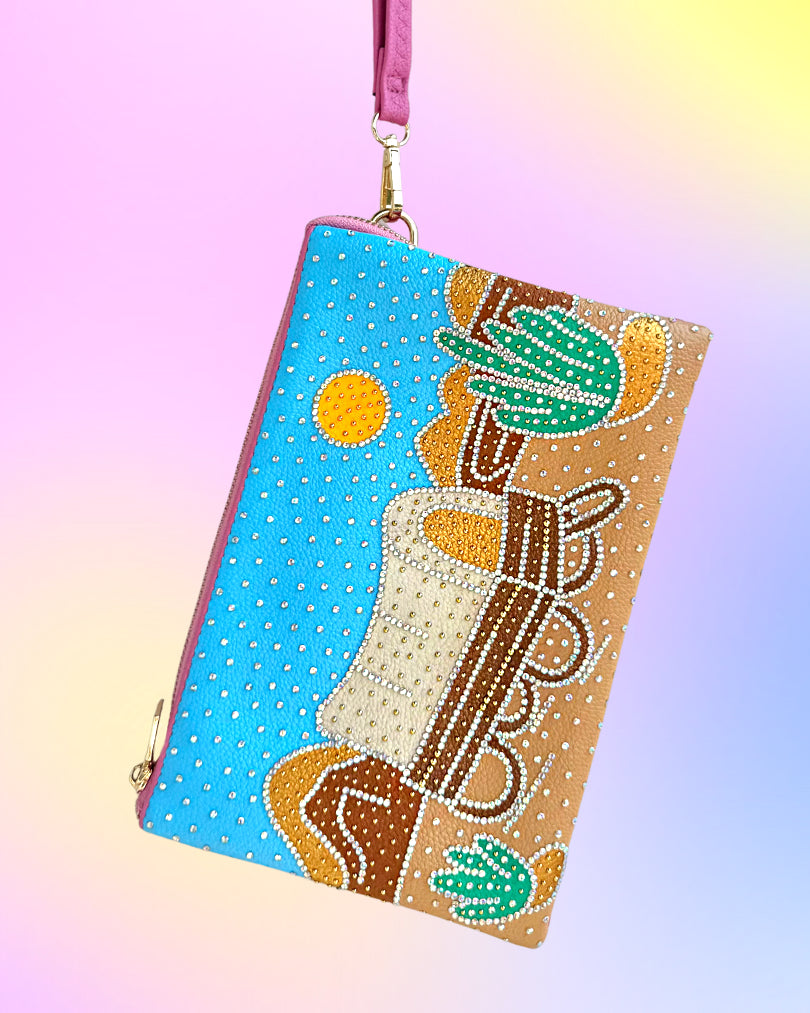 Angled view of desert-scene wristlet with rhinestone embellishments and pink strap.