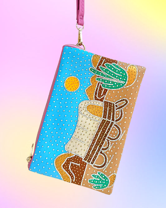 Angled view of desert-scene wristlet with rhinestone embellishments and pink strap.