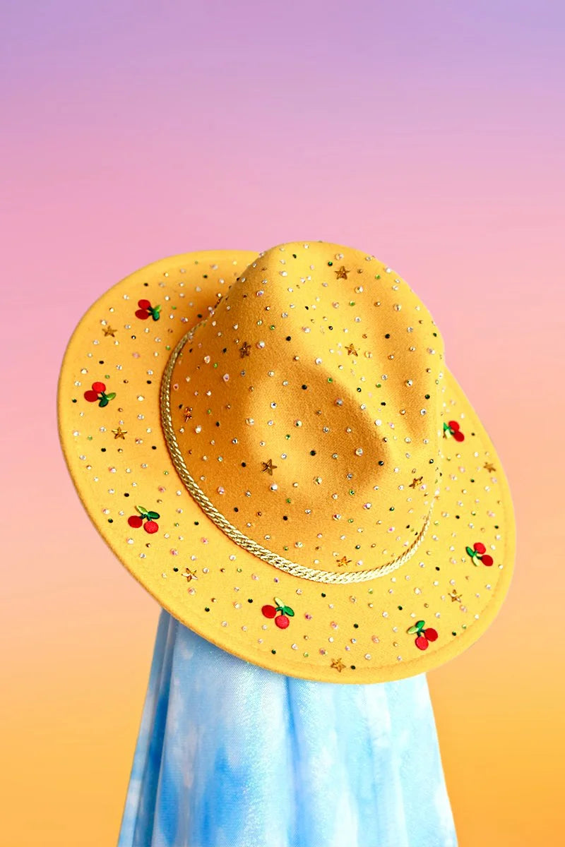 Yellow felt fedora with multicolor rhinestones, gold stars, and embroidered cherry motifs, front angled view.