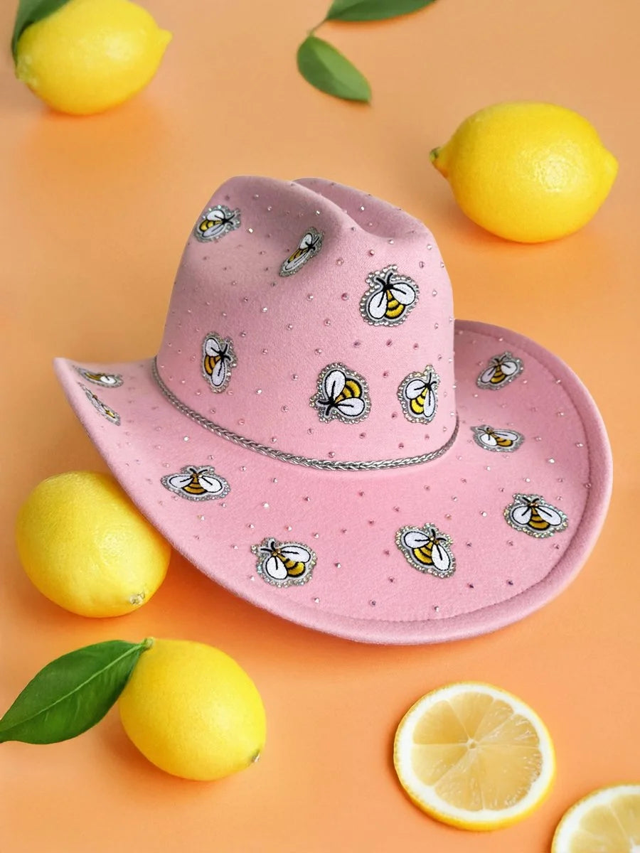 Pink rhinestone cowboy hat with bee motifs styled with lemons on an orange backdrop.