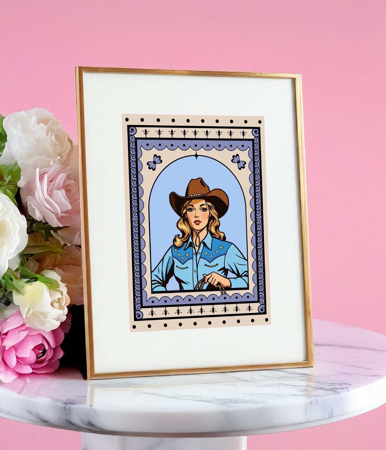Framed cowgirl art print displayed on a round marble stand with pink flowers on a pink background.
