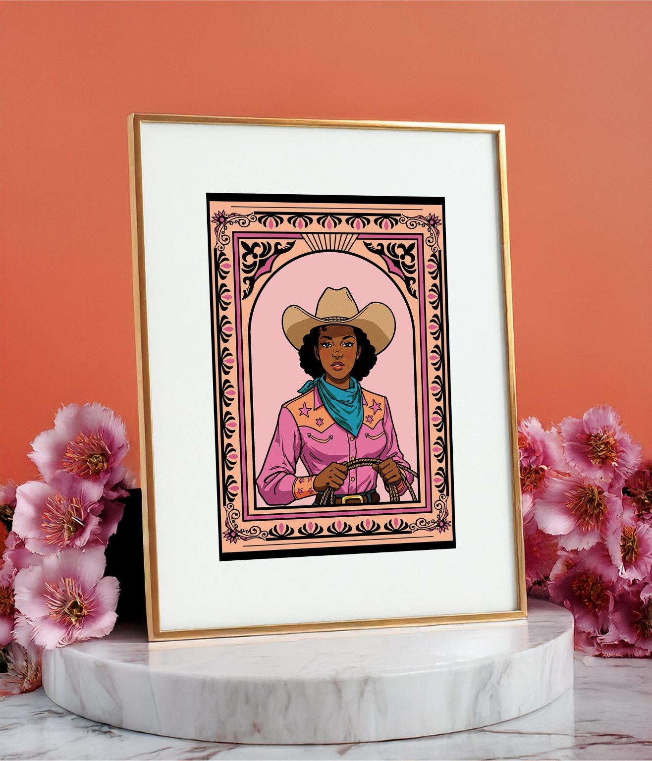 Framed cowgirl art print displayed on a round marble stand with pink flowers on a orange background.