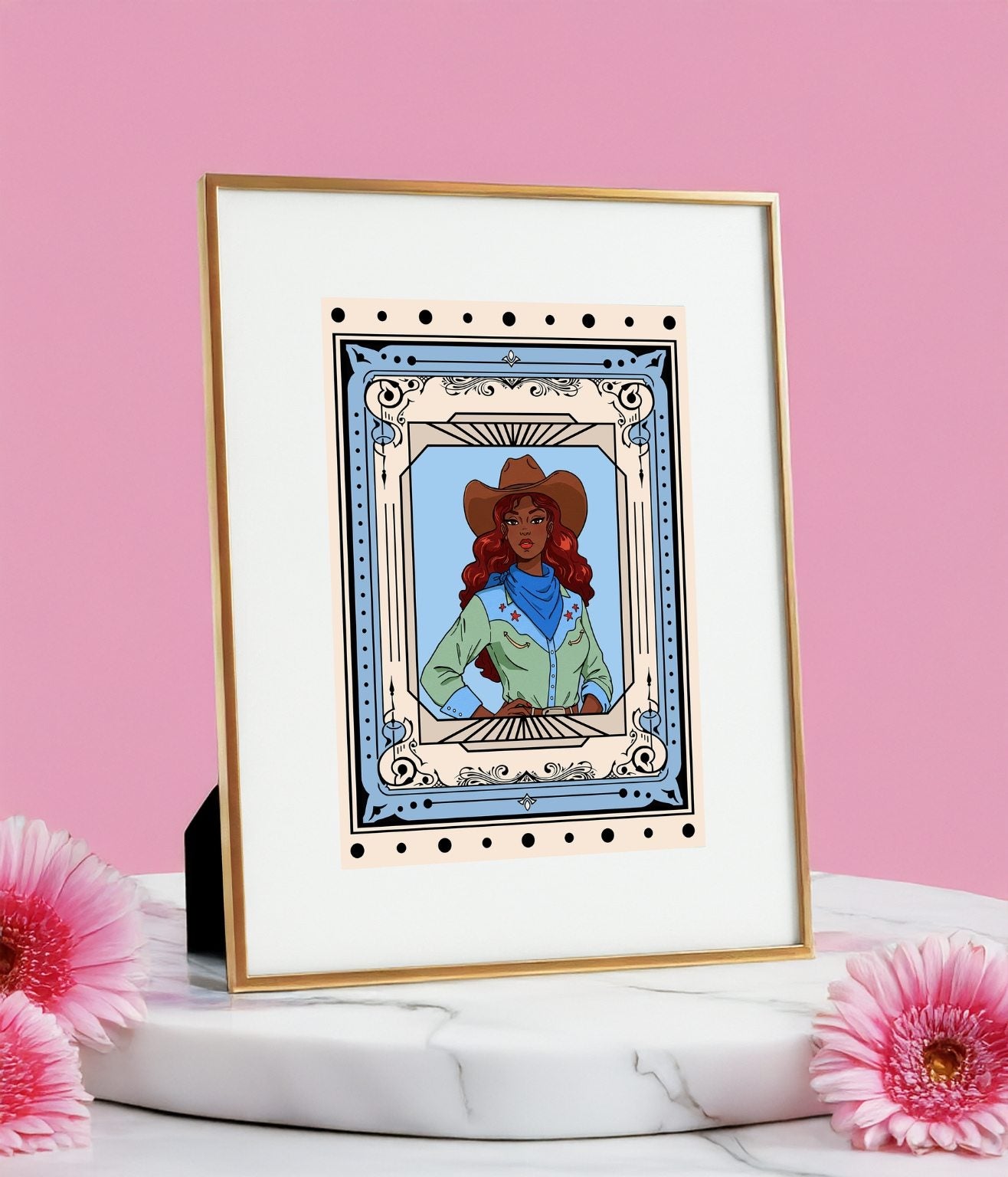 Cowgirl art print in gold frame displayed on a marble stand with pink flowers against a light pink backdrop.