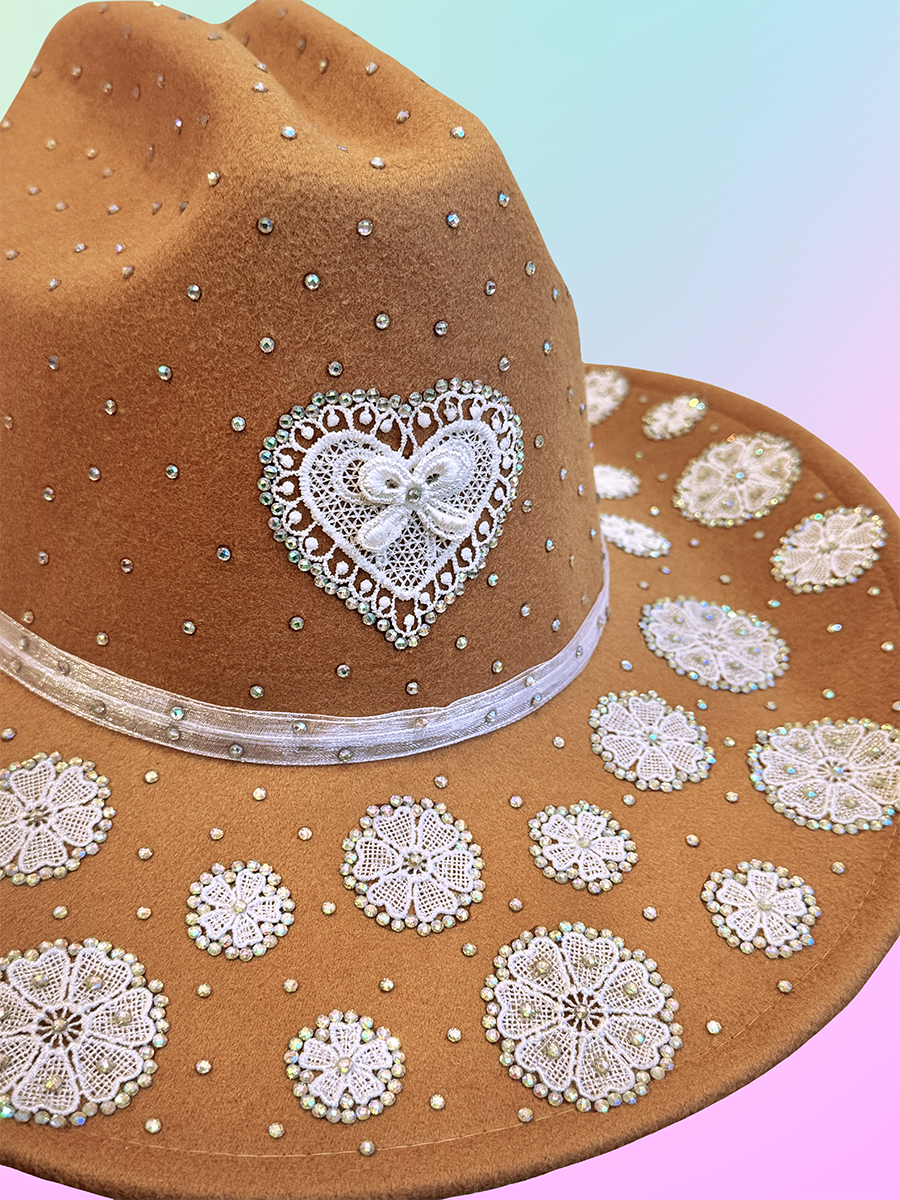 Side angled view of brown lace-decorated cowboy hat featuring white heart applique and shimmering rhinestones.
