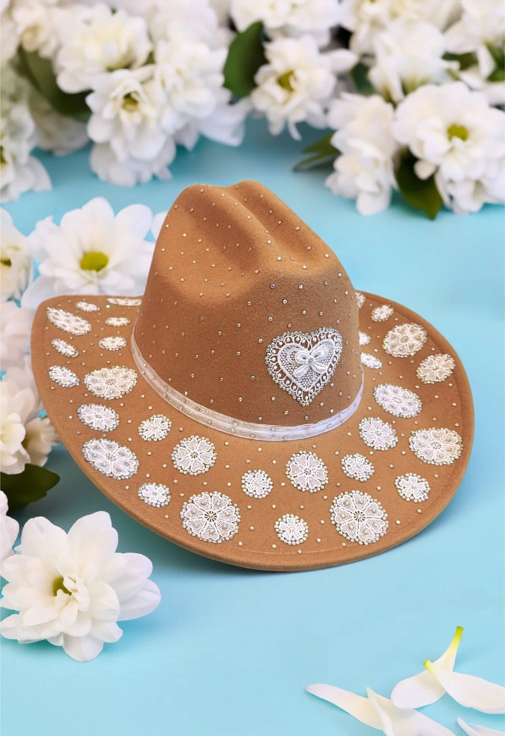 Brown lace-embellished cowboy hat displayed among white flowers on a pastel background.