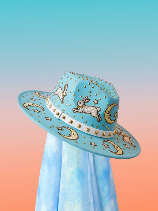 Sky-blue fedora with leaping bunny appliqués, moon and star motifs, and a white studded band, front angled view.