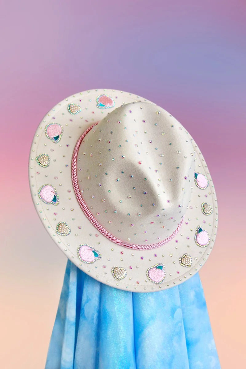 Cream-colored fedora with pink rope band, peach motifs, and scattered rhinestones, front angled view.