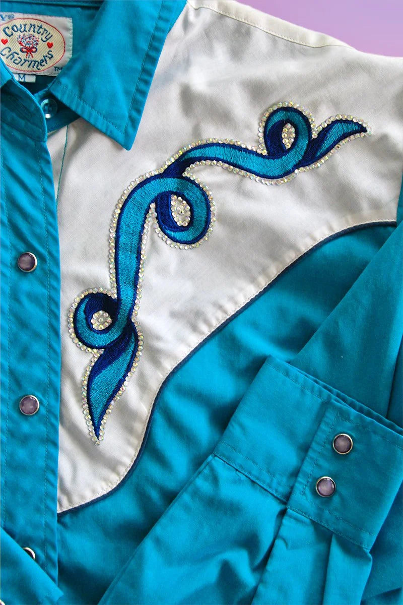 Detailed view of teal and white western shirt with swirling blue embroidery and rhinestone accents.